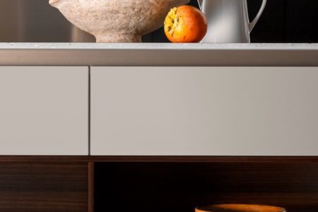 Dettaglio della cucina moderna senza maniglie E0, con ante bianche, una ciotola di legno e frutta su un piano di lavoro minimalista.