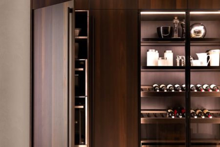 Vista frontale di una parte della cucina senza maniglie moderna E0, con ante in legno scuro e ripiani in vetro illuminati.