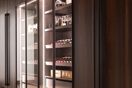 Cucina E0 con gola, armadi in legno scuro e ante in vetro, rivelando scaffali illuminati con bottiglie di vino e accessori da cucina.